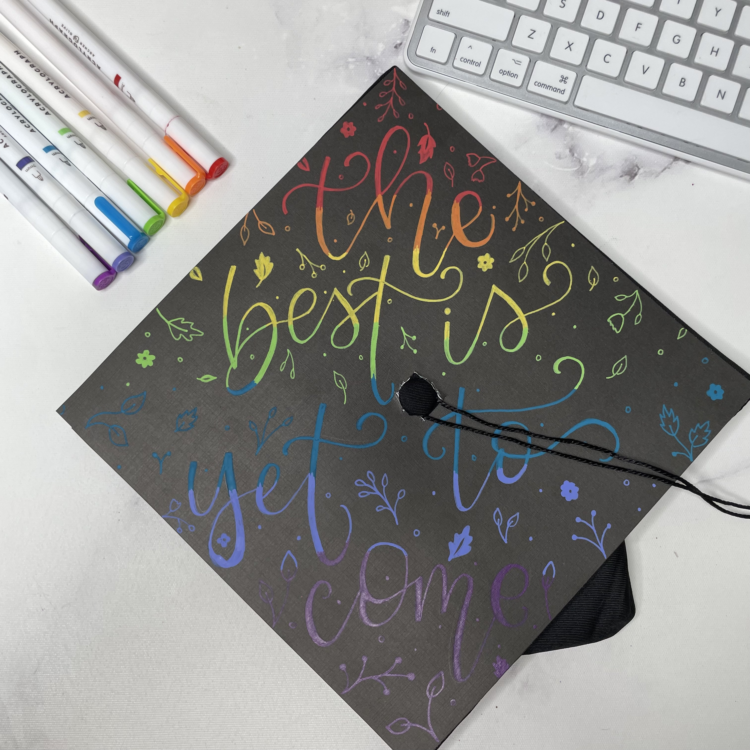 Quote "the best is yet to come" on a black graduation cap in rainbow lettering