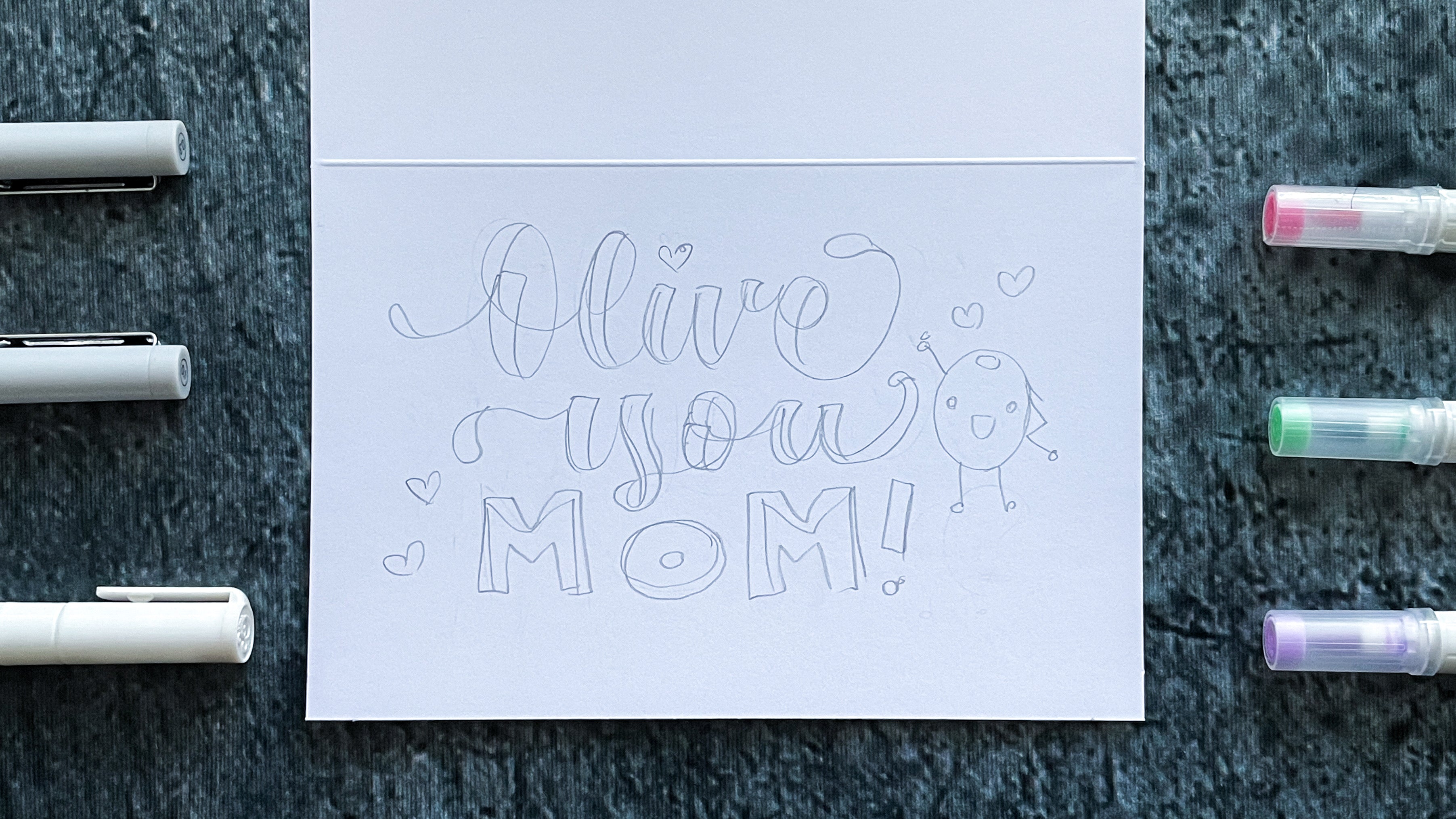 pencil sketch of olive you mom text with kawaii olive doodle and hearts on card stock surrounded by pens