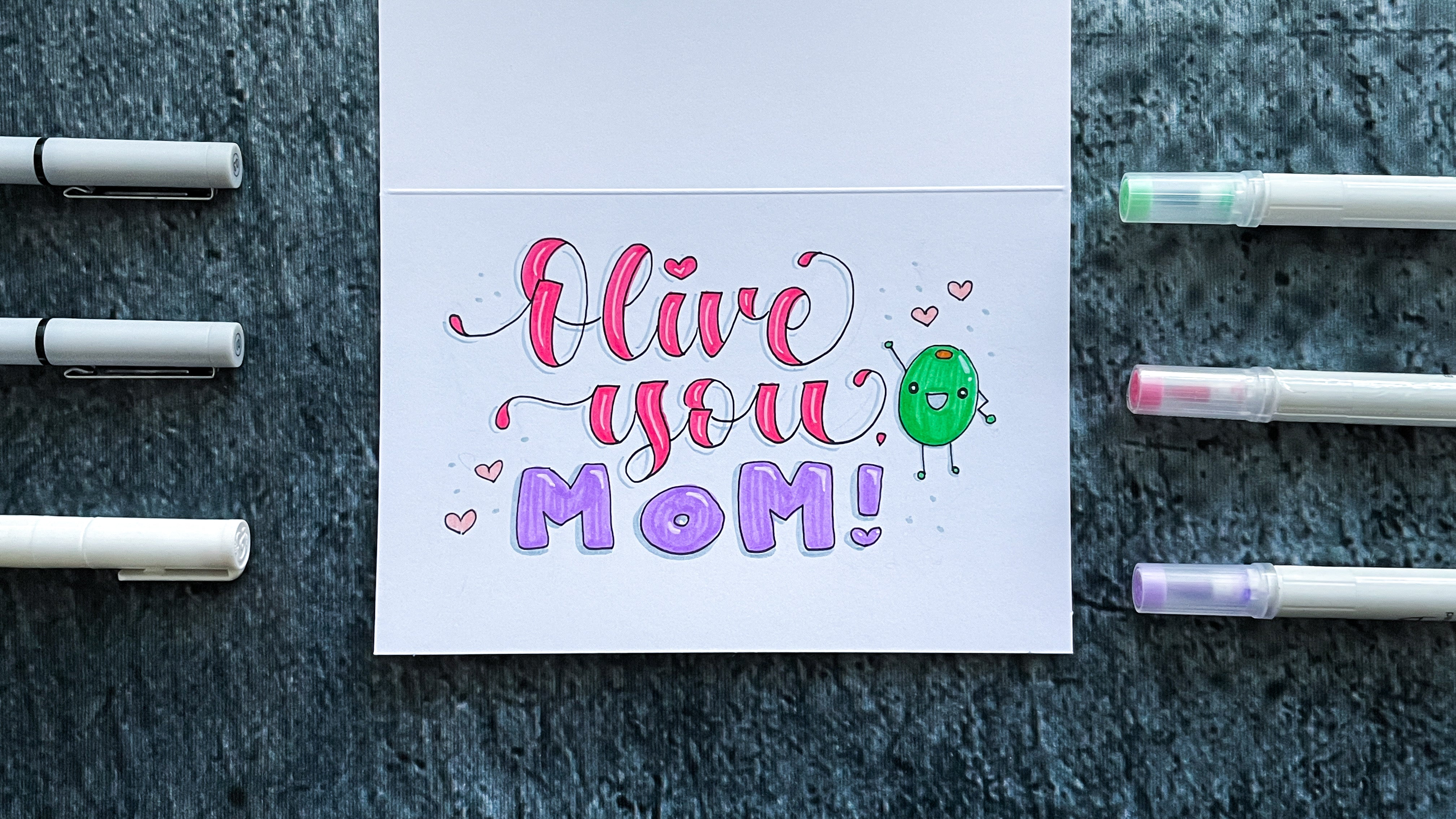Inked and colored text of olive you mom and olive doodle on cardstock surrounded by pens