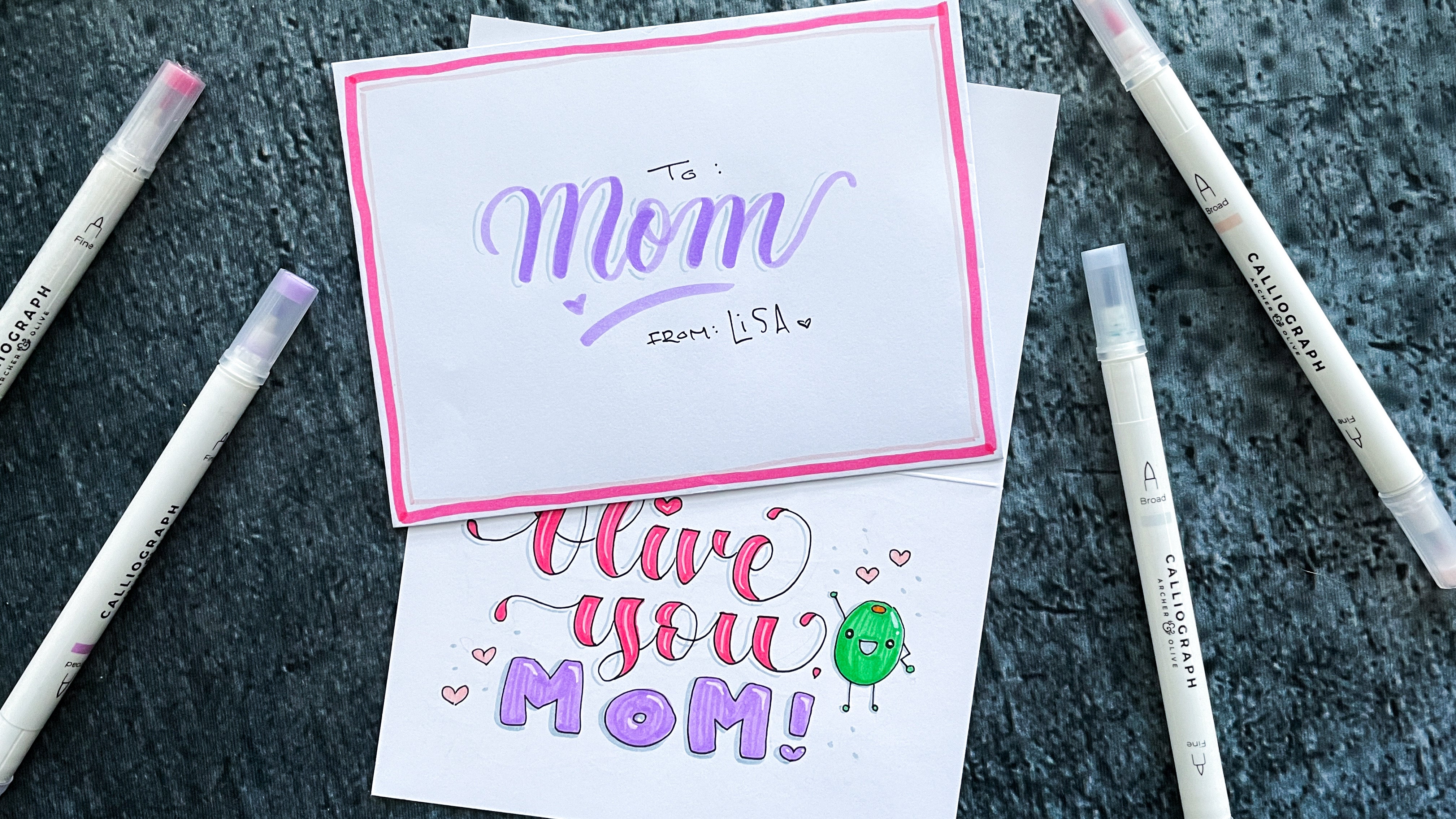 Envelop with mom lettered on it over the completed mother's day card design surrounded by pens