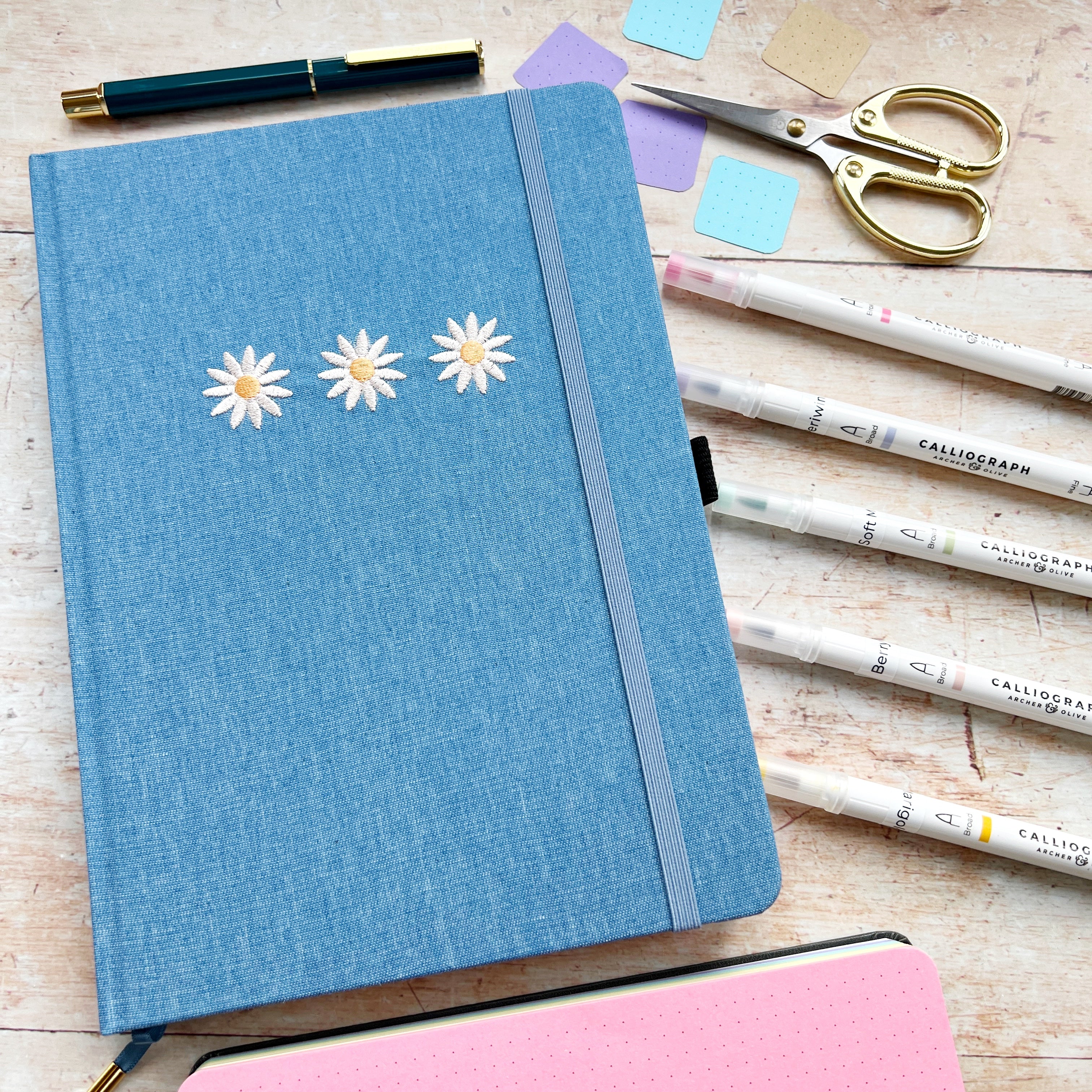 Blue journal with daisies closed and surrounded by pens and accessories