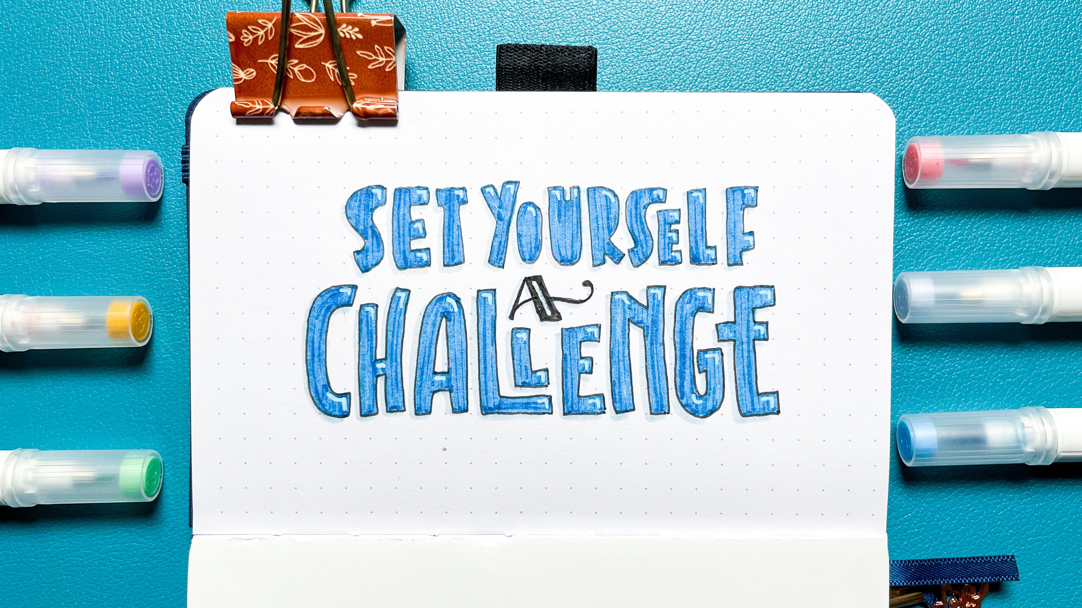 image of hand lettered "set yourself a challenge" in a dot grid journal surrounded by A&O Calliographs