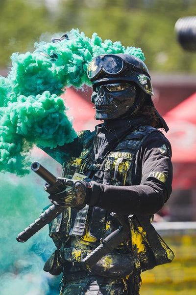 kilt wearing paintballer Reaper stands holding smoke bomb, covered in paint from a battle