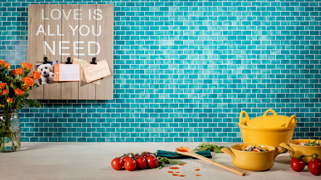 The turquoise small subway tile from the quartz collection used in a kitchen. There is a light tan counter in front of it with yellow pots, tomatoes, and a spoon with tomato sauce on it to the right. To the left there is a clear vase with orange flowers. On the wall is a wooden board with an image of a dog and other little notes pinned to it.