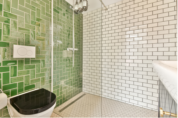 Photo of a tiles shower using green subway tile in a unique pattern, white subway tile on the shower wall, and white hexagon tile on the shower floor. White toilet with black lid. 