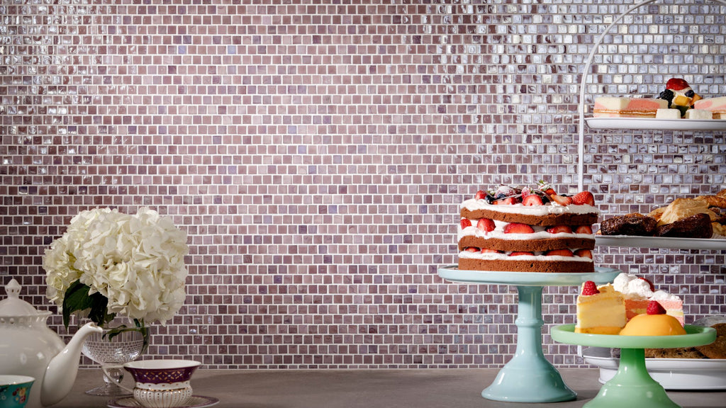 Purple glass tile backsplash in a staggered pattern. In front of it is a display of cakes and foods.