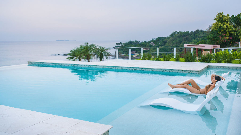 A woman lounging in the Signature Chaise pool lounger on a pool shelf. The pool looks out into the ocean.