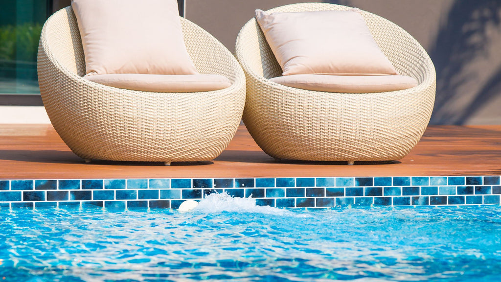 Blue tile from the Vanguard collection installed in a pool waterline. There are 2 modern, round patio chairs sitting on the pool edge.