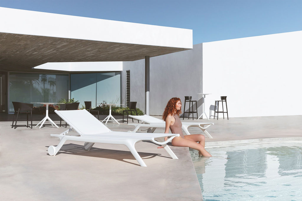 Girl sitting pool side with her legs in the water next to modern patio loungers in white made by Vondom.