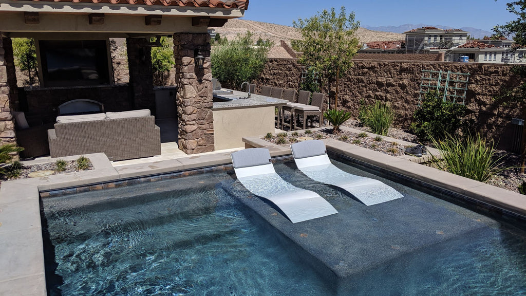 The Curve 2 chaise lounger in the titanium white color with gray headrests. They sit on top of a sun shelf in a pool.
