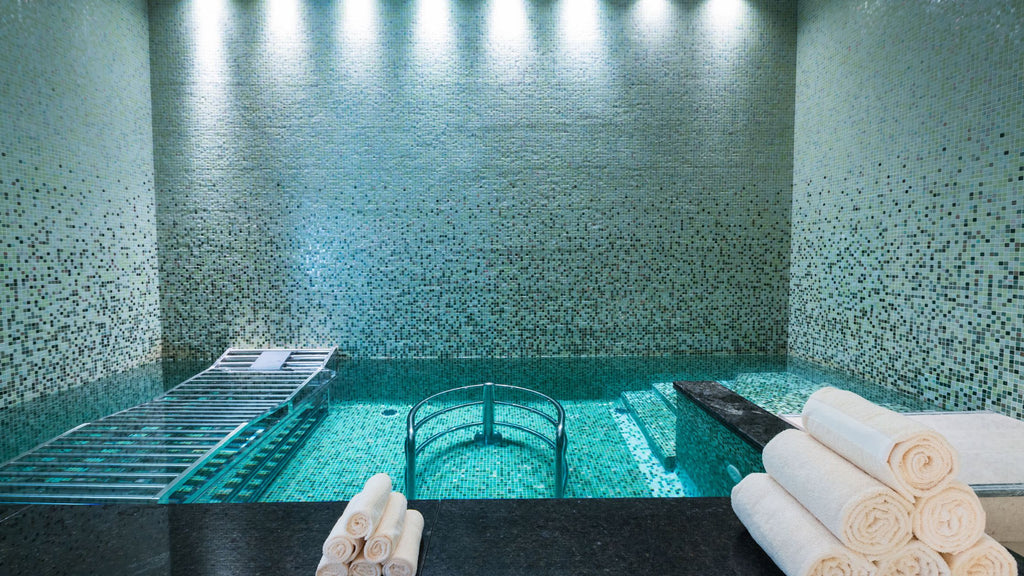 An indoor pool completely tiled in small-format glass tile featuring a gradient from light green and yellow tiles to darker tones and blacks. The walls in the room are completely tiled, blending into the pool directly.