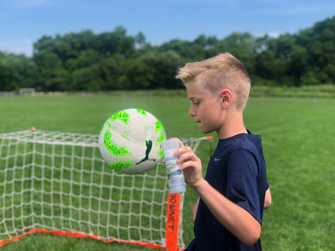Boy playing soccer while holding Dr. Doug's Miracle Balm + Zinc in his hand to help with chafing and blisters from cleats