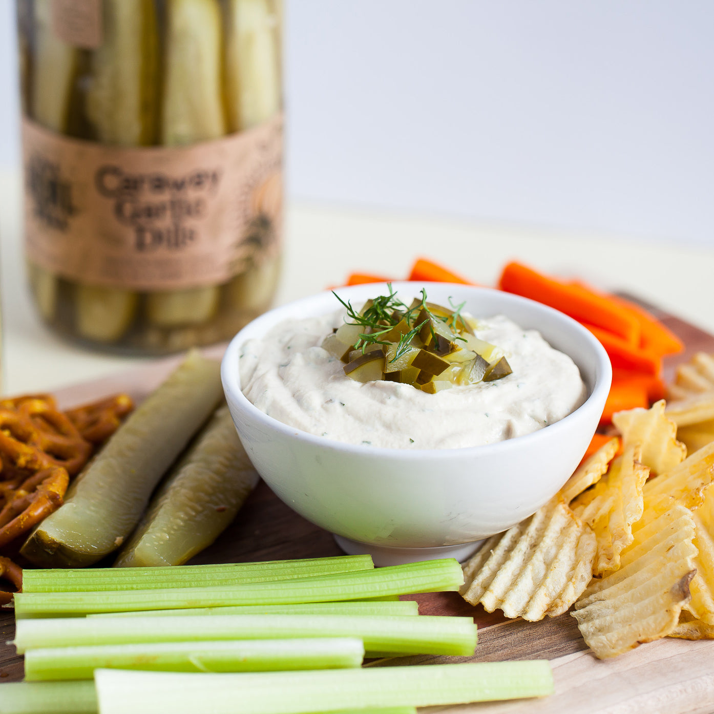 Jar of pickles next to a snack board with pretzels, pickles, celery, potato chips, and vegan dill pickle dip