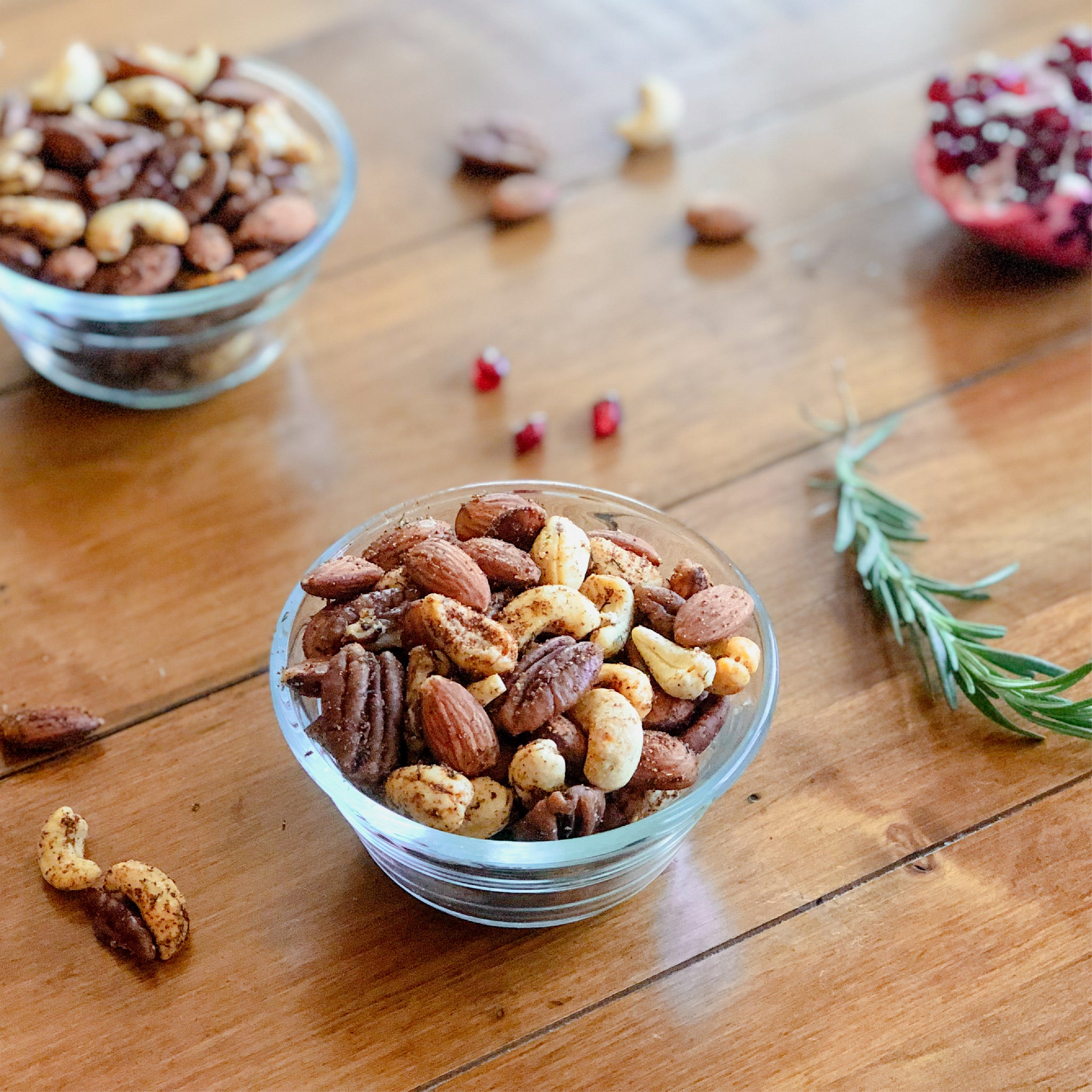 Two bowls of mixed nuts on a table with pomegranate and rosemary