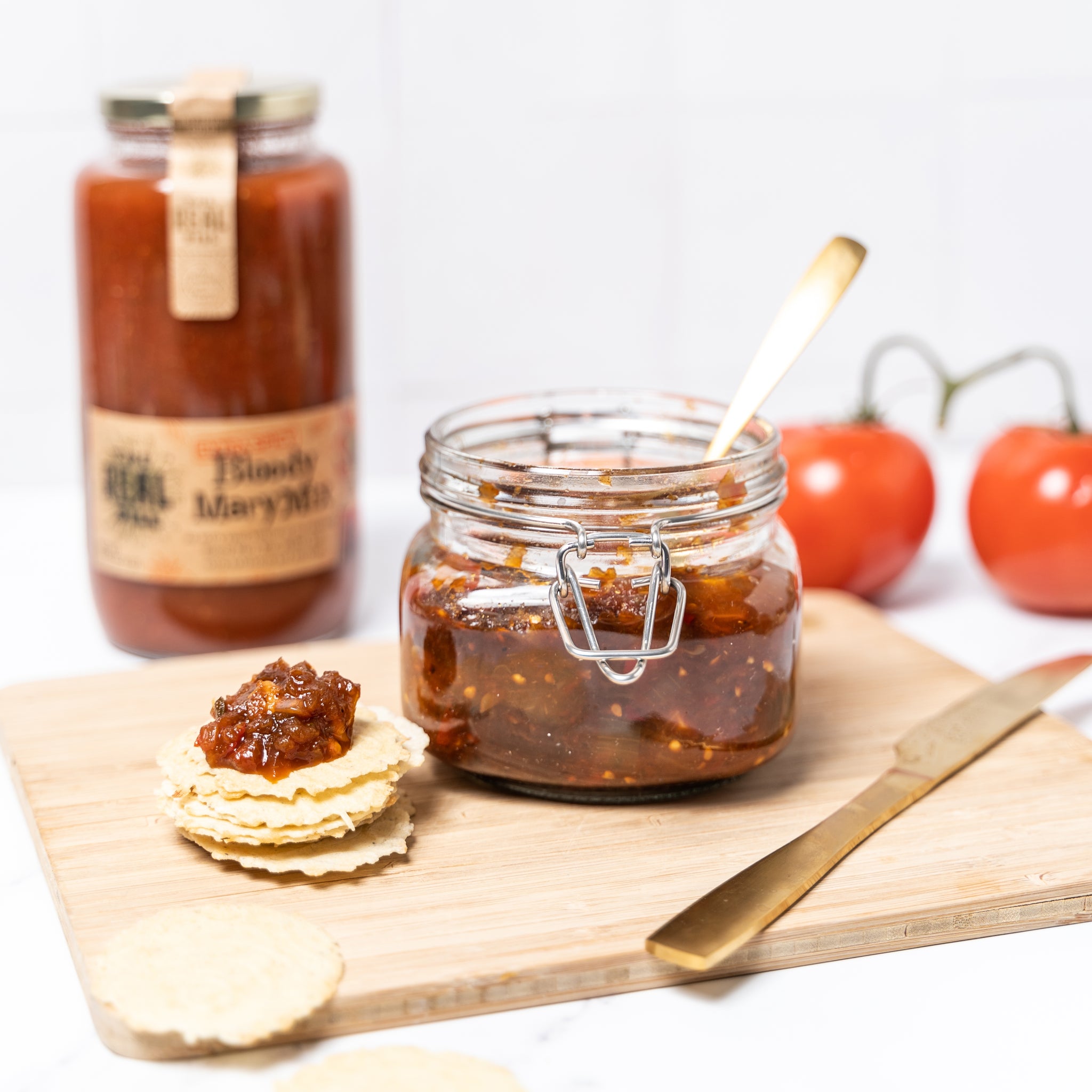 Jar of Extra Spicy Bloody Mary Mix with tomatoes, a wooden board, jar of tomato chutney, crackers topped with chutney, and knife