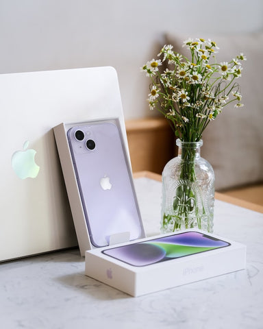 A Purple iPhone 14 in it's box leaning against an Apple MacBook