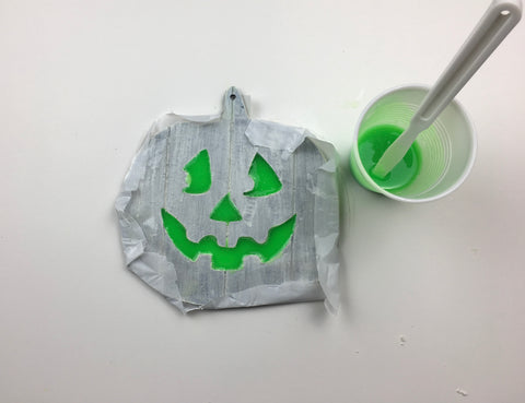 Wooden jack o' lantern sign, with its edges wrapped in wax paper, lying next to a green glow in the dark resin mixture that has also been poured in its eyes and mouth