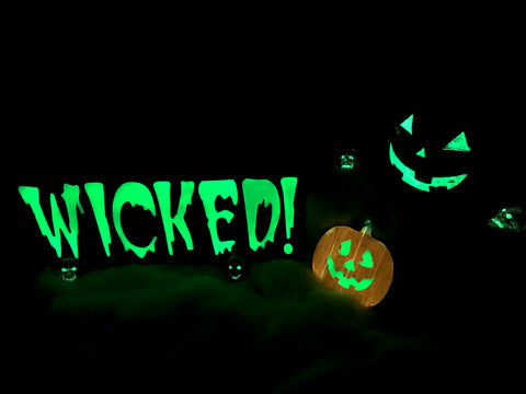 Variety of glow in the dark Halloween decorations placed amongst each other, including 2 glow in the dark jack o' lanterns and a sign that reads "Wicked", as well as several skeletons with glowing eyes and mouths