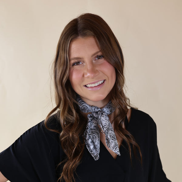 Brunette model wearing a short sleeve, black top with a black and white paisley print tied around her neck. This model is pictured in front of a beige background. 