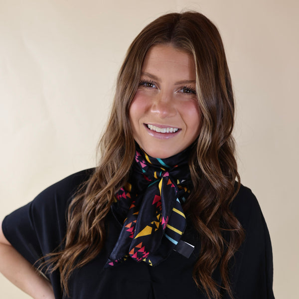 Brunette model wearing a black, drop shoulder top with a black scarf with a multicolored aztec print tied around her neck. Model is pictured in front of a beige background. 