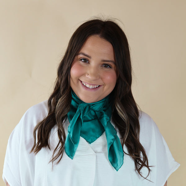 Brunette model wearing a short sleeve, black top with a teal scarf tied around her neck. This model is pictured in front of a beige background. 