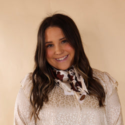 Brunette model wearing an ivory, leopard print top with an ivory, brown, and black cow print scarf tied around her neck. Model is pictured in front of a beige background. 