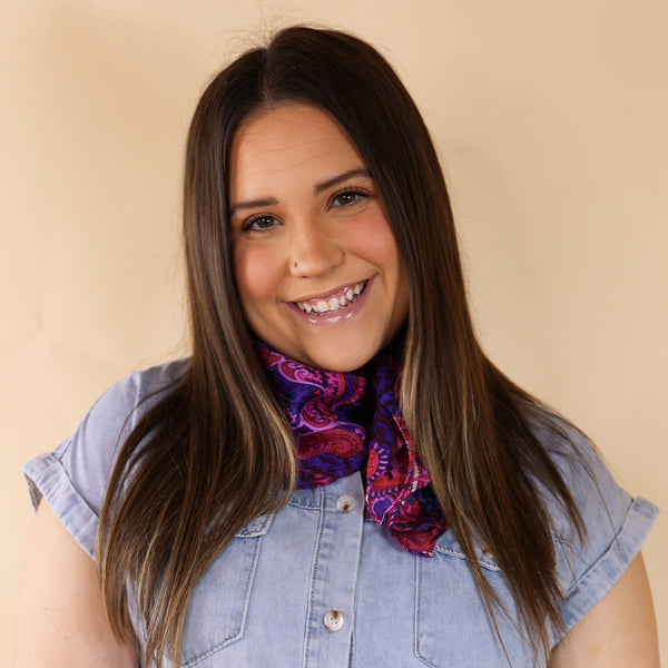 Brunette model is pictured wearing a denim button up top and a purple and pink mix paisley printed scarf tied around her neck. She is pictured in front of a beige background. 