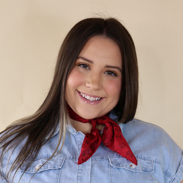 Brunette model is pictured wearing a denim button up top and a red scarf with a black print tied around her neck. She is pictured in front of a beige background. 