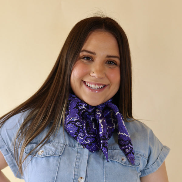 Brunette model is pictured wearing a denim button up top and a purple and black mix paisley printed scarf tied around her neck. She is pictured in front of a beige background. 
