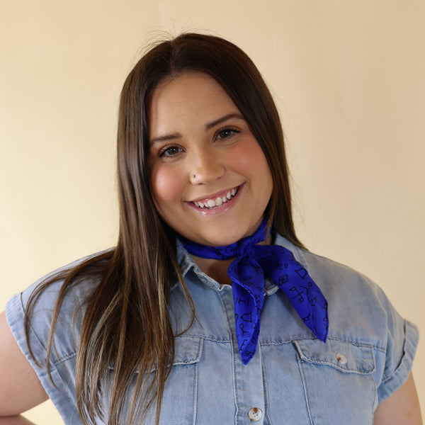 Brunette model wearing a blue dress with Royal Blue Branded print scarf tied around her neck. Model is pictured in front of a beige background.