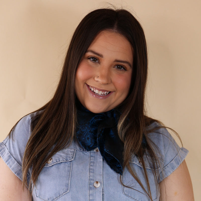 Brunette model is wearing a blue dress with a blue scarf tied around her neck. Model is standing in front of beige background.