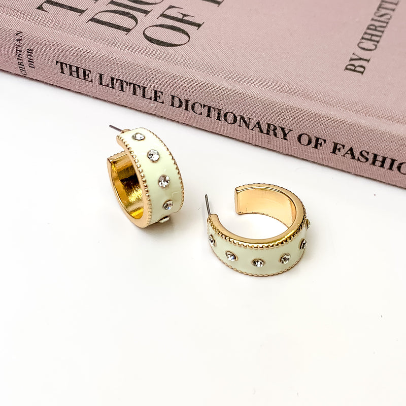 Thick, gold hoop earrings with a ivory inlay on the outside. These hoops also include a single line of clear crystals through the middle. One hoop is pictured stadning up right and the other is pictured laying on its side. These hoops are pictured on a white background with a mauve colored book above the earrings. 