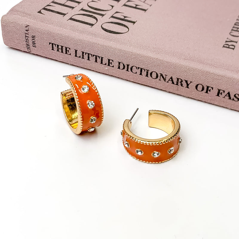 Thick, gold hoop earrings with a cognac brown inlay on the outside. These hoops also include a single line of clear crystals through the middle. One hoop is pictured stadning up right and the other is pictured laying on its side. These hoops are pictured on a white background with a mauve colored book above the earrings. 