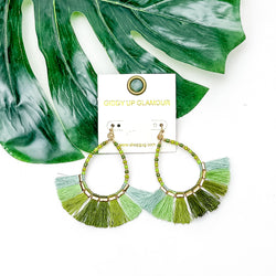 Beaded Open Teardrop Earrings With Fringe Bottom in Green Tones. Pictured on a white background with a leaf behind the earrings.