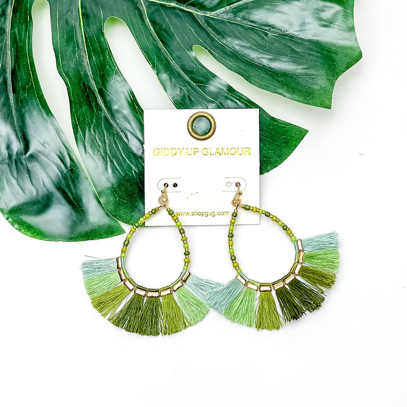 Beaded Open Teardrop Earrings With Fringe Bottom in Green Tones. Pictured on a white background with a leaf behind the earrings.