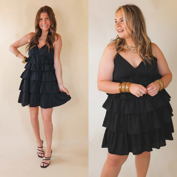 A short black dress featuring a V neckline, spaghetti straps, and a tiered ruffle skirt. Item is pictured on a pale pink background.