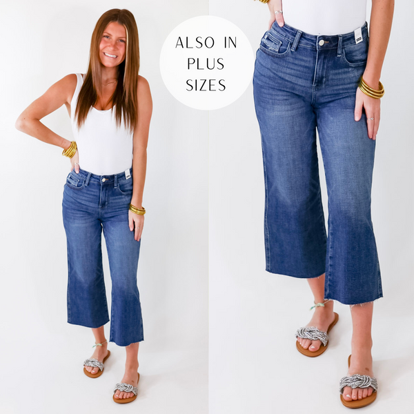 Model is wearing a pair of wide leg cropped jeans in dark wash. Model has it paired with a white tank top, crystal sandals, and gold jewelry.
