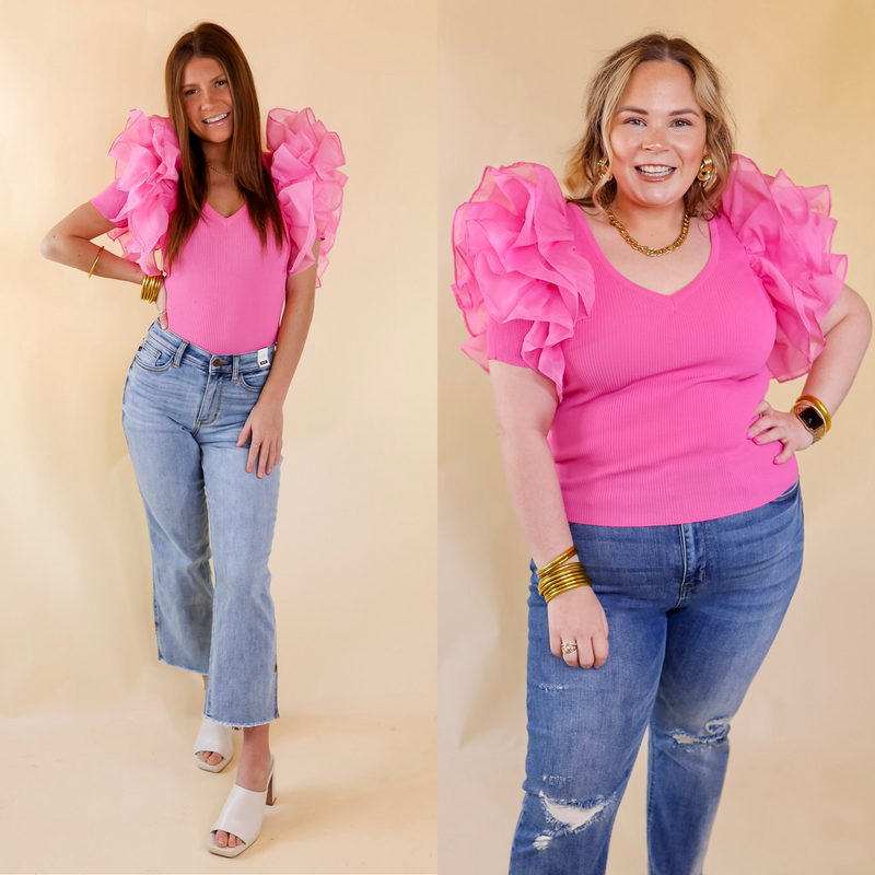 Model is wearing a pink fitted top with large ruffle sleeves. Model has this top paired with distressed jeans, white heels, and gold jewelry.