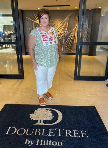 Plus size woman wearing white jeans, tan sandals, and a green leopard print top with a pink and red embroidered yoke stands at the entrance of the Doubletree by Hilton smiling at the camera. 