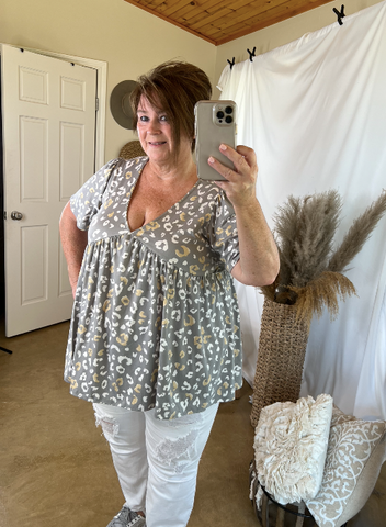 plus size woman stands in front of a mirror taking a selfie while wearing white jeans and a grey v-neck leopard print top. In the background is a jute run and pampas grass. 