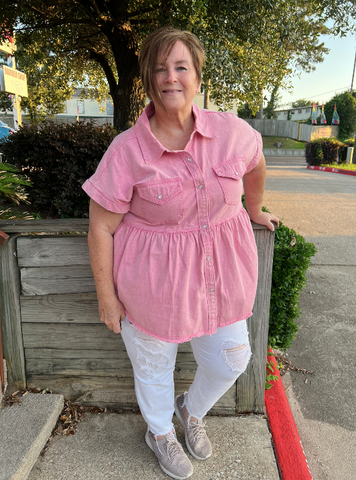 Plus size woman in white jeans, grey sandals, and a pink, short sleeved, pearl button down top stands in front of a garden planter smiling at the camera. 