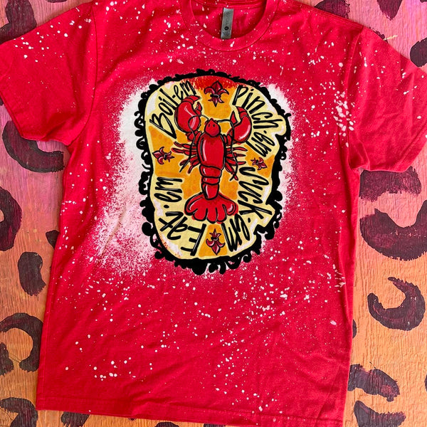 A red short sleeve shirt with a bleach splatter design and a large bleach spot in the center featuring a graphic of a red crawfish with an asymmetric orange circle behind it. The words "Boil-em, pinch-em, suck-em, eat-em" circle the crawfish and a black border can be found around the orange background.