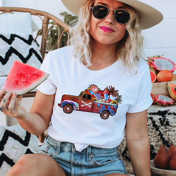 A white short sleeve shirt with a graphic of a red, white, brown, and blue truck carrying fourth of july decorations and fireworks. 