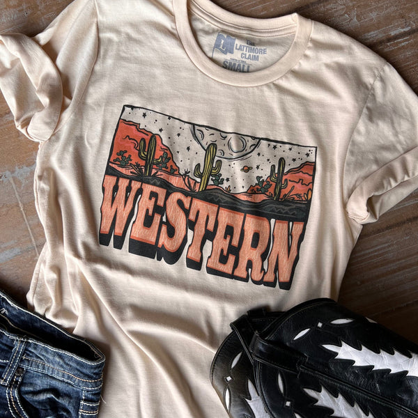 A white shirt featuring a desert at night with stars, a moon, cacti, and cliffs. The words "western" can be found right below the graphic. Text is light orange with a slight wave. Item is pictured on a wood background with black boots and denim shorts.