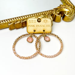 Circle drop earrings with a hanging flamingo ignite cushion cut crystal. The pendant is half gold beads and half light pink crystal beads. These earrings are pictured on a wood earrings holder in front of a gold mirror on a white background. 
