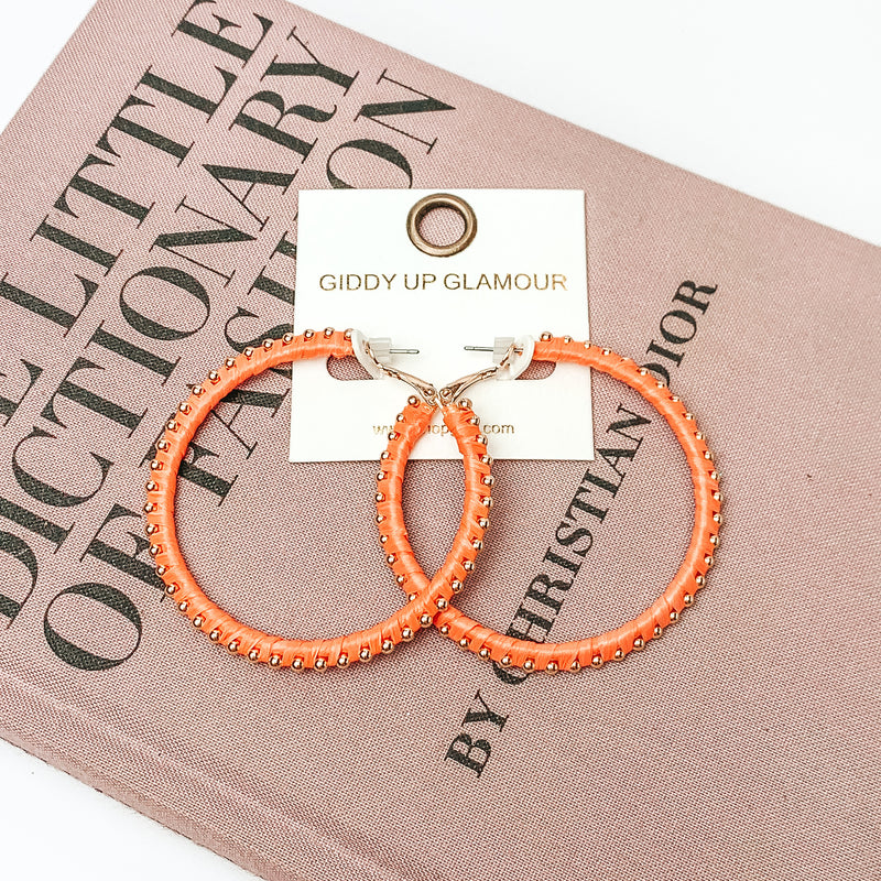 Pictured are circle orange hoop earrings with gold beads around it. They are pictured with a pink fashion journal on a white background.