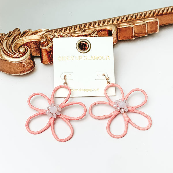 Pictured are gold fish hook earrings with a flower outline pendant. This pendant is light pink and has center light pink crystals. These earrings are pictured in front of a gold mirror on a white background. 