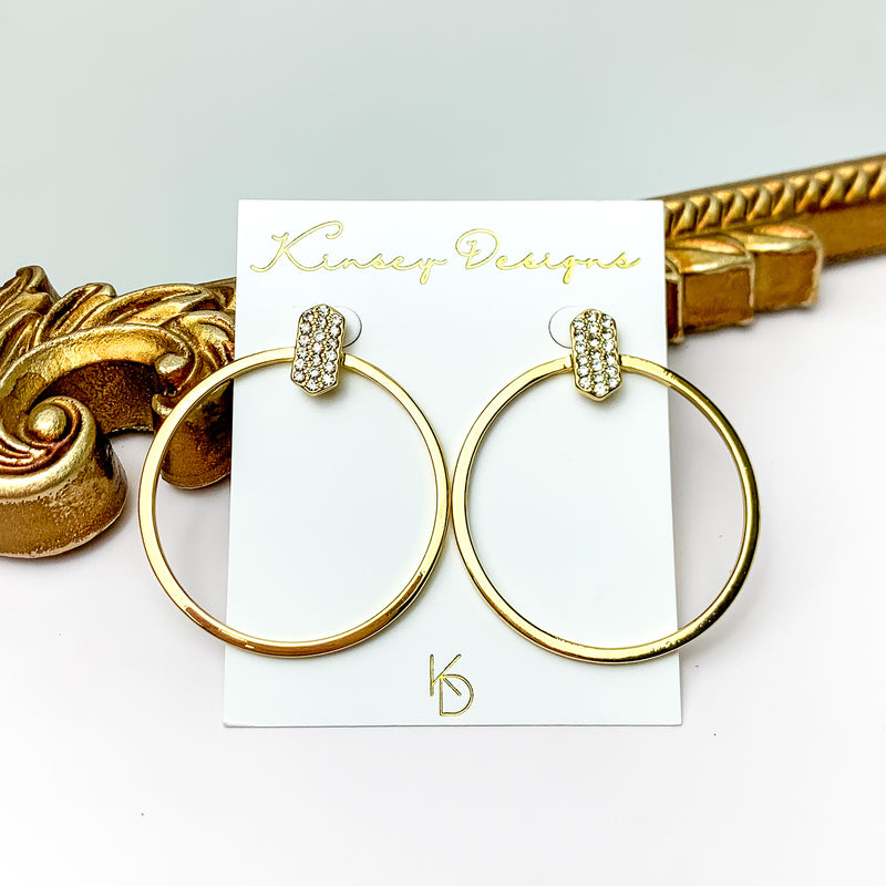 A pair of gold hoop earrings with an oval bar post back encrusted in clear crystals. These earrings are pictured in front of a gold mirror on a white background.  
