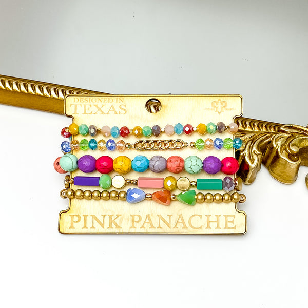 Set of five multi crystal, multi beads, and gold bead bracelets in different sizes. These braceets are pictured on a Pink Panache wood holder in front of a gold mirror and on a white background. 
