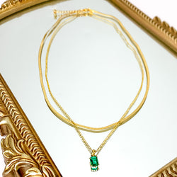 Pictured is double layered chain necklace with a single, green crystal pendant. This necklace is pictured on a white background with a gold mirror in the top right corner.  
